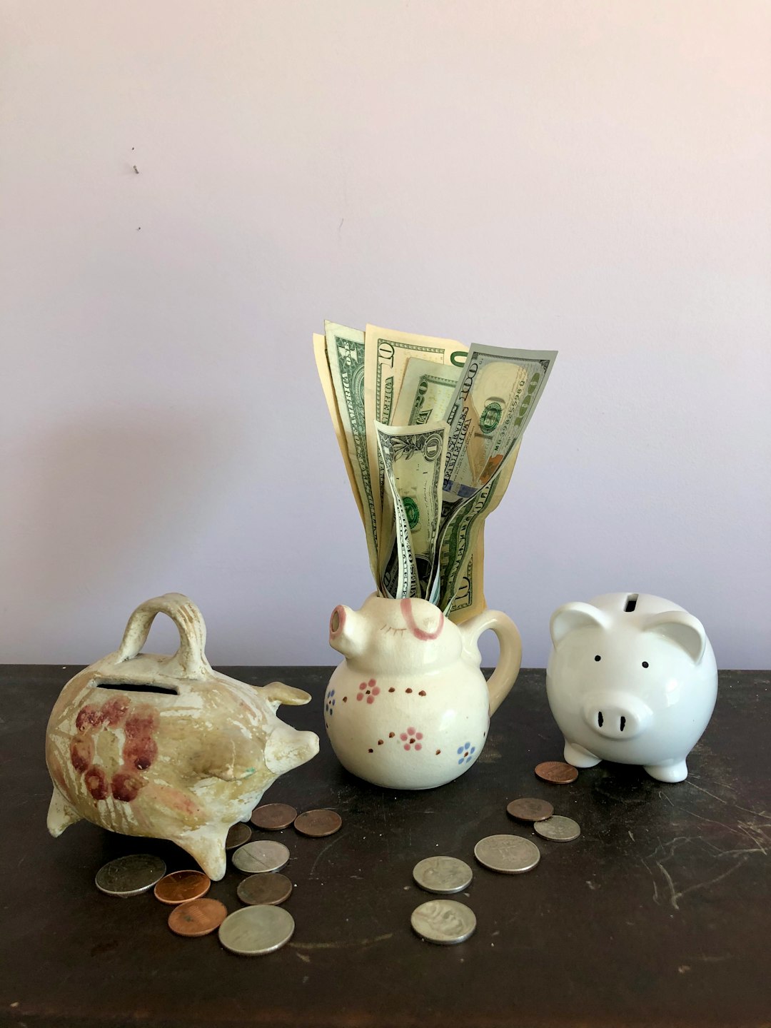 Two ceramic piggy banks and a small ceramic pig pitcher in a row on a wooden cabinet against a light lavender grey wall. US coins are scattered on the cabinet. A cluster of US paper currency in a variety of denominations sprout from the pig pitcher.